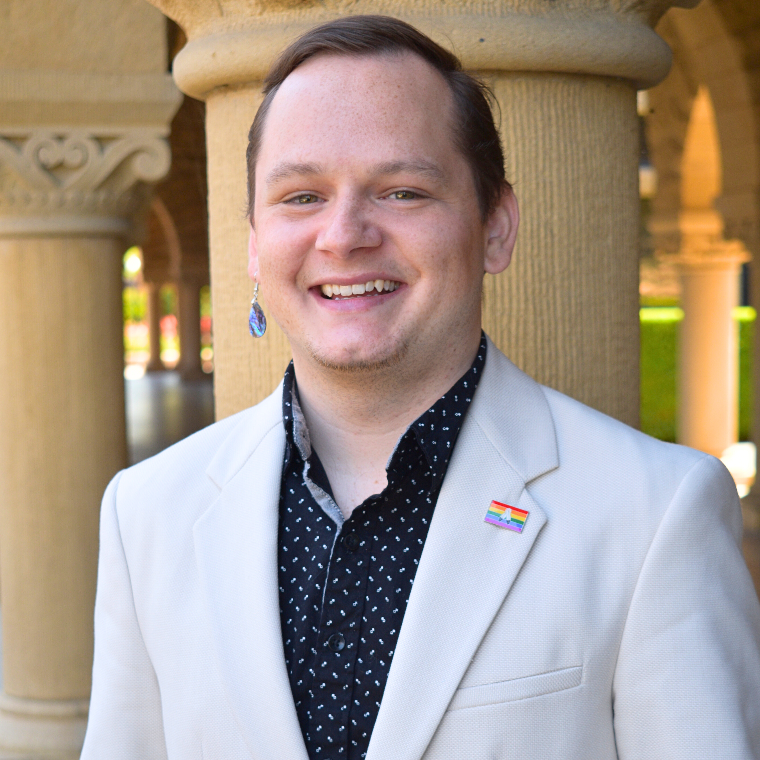 Preston is pictured in foreground smiling in black-and-white patterned shirt with beige suit jacket. The columns of Stanford University's arcades on a sunny day are in the background.